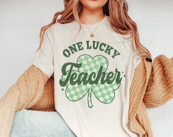 Puede incluir: Una camiseta blanca con un diseño de trébol verde y el texto "One Lucky Teacher" en una fuente retro.