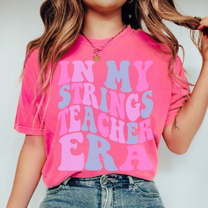 May include: Pink t-shirt with the text "In My Strings Teacher Era" in a retro font.