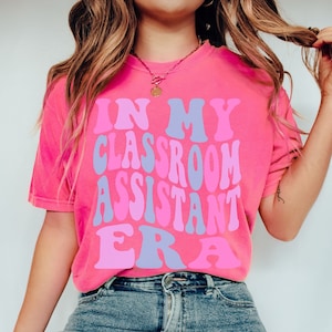 May include: Pink t-shirt with the text "In My Classroom Assistant Era" in a retro font with pink, blue, and purple colors.