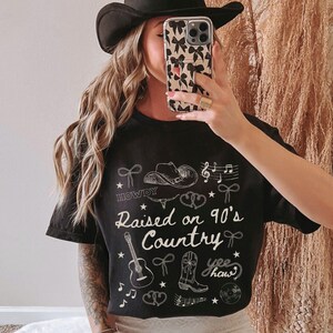 May include: Black t-shirt with white graphic text and illustrations. The text reads "Raised on 90's Country" with cowboy-themed drawings. The shirt also features the word "Howdy" and the phrase "yee haw".