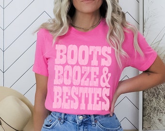 May include: A pink t-shirt with the text "Boots Booze & Besties" in white. The shirt is being worn by a person and is available in size small.