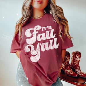 May include: Brick-colored t-shirt with the text "It's Fall Y'all" in a retro font. The shirt is paired with gray shorts and cowboy boots. The boots are brown with decorative stitching.