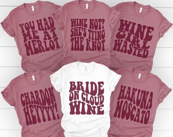 May include: Six t-shirts with different wine-themed phrases printed in a retro font. The shirts are in shades of maroon and white. The phrases include "You Had Me at Merlot", "Wine Not? She's Tying the Knot", "Wine Girl Wasted", "Chardon Heyyyy!", "Bride on Cloud Wine", and "Hakuna Moscato".