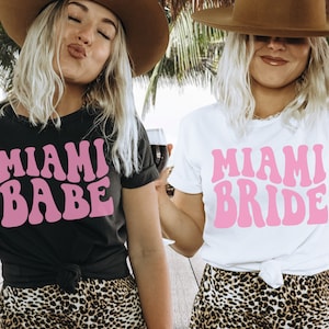 Pode incluir: Duas mulheres vestindo camisetas combinando com o texto "Miami Babe" e "Miami Bride" em fonte retr&ocirc; rosa. As mulheres est&atilde;o usando chap&eacute;us e sorrindo.