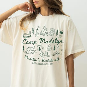 May include: Ivory-colored t-shirt with the text "Camp Madelyn" in moss green, surrounded by camping-themed illustrations. The text "Madelyn's Bachelorette" and "Breckenridge, CO" are also printed on the shirt.