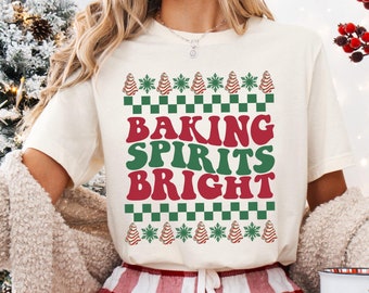 May include: A white t-shirt with a red and green Christmas-themed design that says "Baking Spirits Bright". The shirt is being worn by a person with red and white plaid pants.