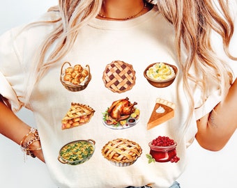 Può includere: Una maglietta bianca a tema Ringraziamento con illustrazioni di vari cibi tra cui torta, purè di patate, tacchino, salsa di mirtilli rossi e panini. Il testo "size small IVORY" è visibile nella parte inferiore dell'immagine.