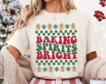 May include: A white t-shirt with a red, green, and white design that says "Baking Spirits Bright". The design features gingerbread men and snowflakes. The shirt is being worn by a person wearing red and white plaid pants. The word "NATURAL" is written in white letters on a black background at the bottom of the image.