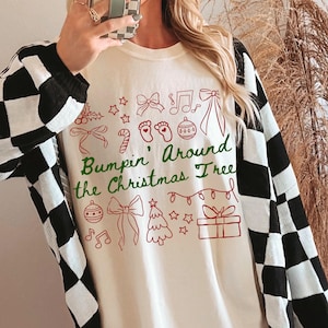 May include: An ivory-coloured long-sleeved shirt with the text "Bumpin' Around the Christmas Tree" in green and red, featuring Christmas doodles. A black and white chequered cardigan is worn over the shirt. The size is L.