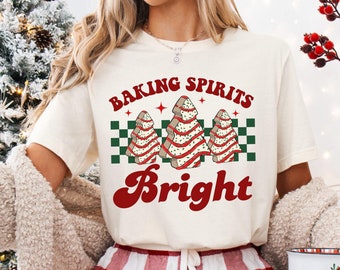 May include: A white t-shirt with the text "Baking Spirits Bright" in red and black lettering. The design features three stylized Christmas trees with red and green frosting.