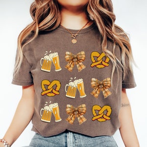 May include: A brown t-shirt with a graphic design of repeating images of pretzels, beer steins, and plaid bows.