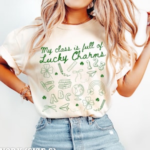 May include: Ivory t-shirt with the text "My class is full of Lucky Charms" in green lettering. The shirt features a variety of green shamrock and school-themed illustrations.