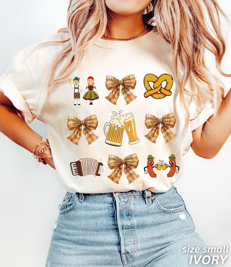 Op de afbeelding: Een ivoorwit T-shirt met een bruine en gele geruite strik, een pretzel, een bierpul, een paar worstjes, een accordeon en een stel in traditionele Duitse kleding. De tekst "size small IVORY" staat onderaan de afbeelding.