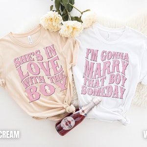 Puede incluir: Dos camisetas con texto rosa sobre fondo blanco. La camiseta de la izquierda dice "She's in love with the boy" en rosa sobre un fondo crema suave. La camiseta de la derecha dice "I'm gonna marry that boy someday" en rosa sobre un fondo blanco.