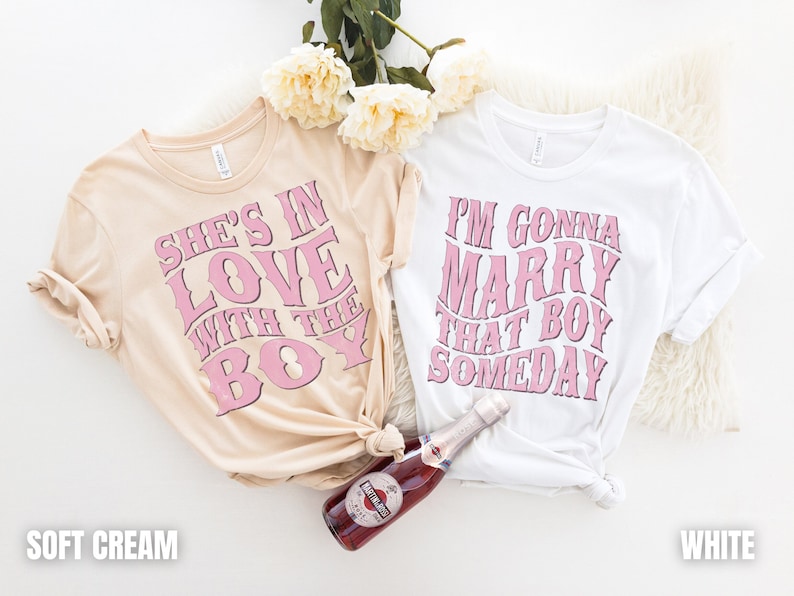 Puede incluir: Dos camisetas con texto rosa sobre fondo blanco. La camiseta de la izquierda dice "She's in love with the boy" en rosa sobre un fondo crema suave. La camiseta de la derecha dice "I'm gonna marry that boy someday" en rosa sobre un fondo blanco.