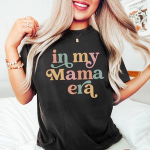 Op de afbeelding: Een zwart T-shirt met de tekst "in my mama era" in kleurrijke, gestileerde letters.