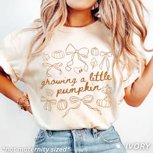 May include: Cream-coloured t-shirt with the phrase "growing a little pumpkin" in brown script. The shirt features brown line art of pumpkins, bows, and leaves. The text "*not maternity sized*" is at the bottom.
