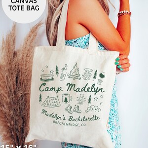 May include: Natural canvas tote bag, 15" x 16", with a green design on one side. The design includes camping-themed illustrations and the text "Camp Madelyn" and "Madelyn's Bachelorette, Breckenridge, CO". Made of 100% cotton.
