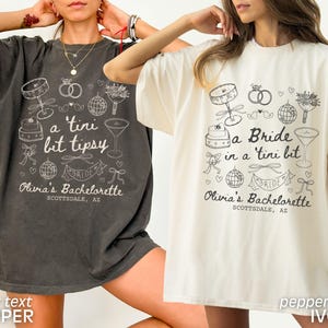 Op de afbeelding: Twee oversized t-shirts met een vrijgezellenfeestthema. De shirts bevatten een bruid, een cocktailglas, een taart en andere bruiloftgerelateerde afbeeldingen. De tekst op de shirts luidt "a tini bit tipsy" en "a Bride in a tini bit". De tekst op de shirts is in ivoor en de shirts zijn in peper en ivoor.