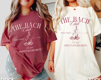 Puede incluir: Dos camisetas de gran tamaño con un gráfico de temática vinícola que dice "The Bach Club" con el texto "HAPA VALLEY - CA" y "Est. 2023 ASHLEY'S BACHELORETTE". La camiseta de la izquierda es de color rojo ladrillo y la camiseta de la derecha es de color marfil.