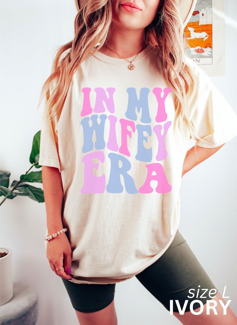 May include: A white t-shirt with the text "In My Wife Era" in pink and blue retro-style lettering. The shirt is size L and is ivory in color.