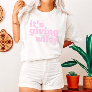 May include: White t-shirt with pink text that reads "it's giving wifey".