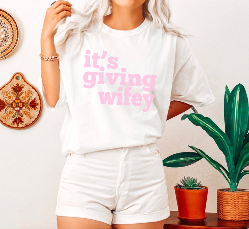 May include: White t-shirt with pink text that reads "it's giving wifey".