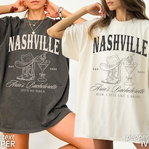 May include: Two oversized t-shirts, one dark gray and one cream, featuring a Nashville-themed graphic. The design includes a cowboy hat, boots, and a cocktail glass, with the text "Nashville", "Aria's Bachelorette", and "Let's Go Girls" or "Mah, I Feel Like a Bride".