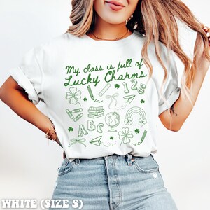 May include: White t-shirt with green text that reads "My class is full of Lucky Charms" and green illustrations of school supplies, including a pencil, ruler, paper airplane, globe, books, and four-leaf clovers.