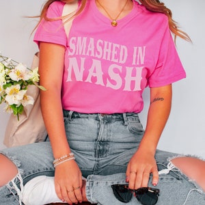 K&ouml;nnte beinhalten: Ein leuchtend pinkfarbenes T-Shirt mit dem Text "Smashed in Nash" in hellrosa, im Vintage-Stil. Das Shirt wird mit hellblauen, zerrissenen Jeans kombiniert. Accessoires sind eine goldene Halskette, Armb&auml;nder und Sonnenbrillen.