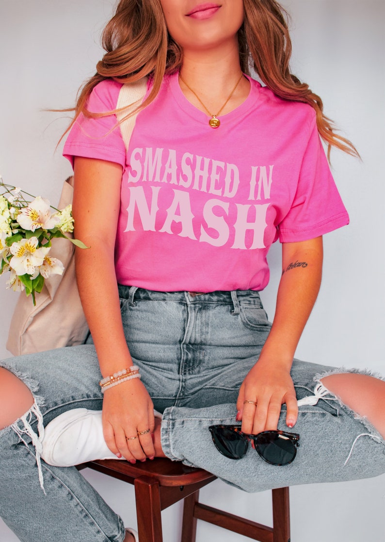 K&ouml;nnte beinhalten: Ein leuchtend pinkfarbenes T-Shirt mit dem Text "Smashed in Nash" in hellrosa, im Vintage-Stil. Das Shirt wird mit hellblauen, zerrissenen Jeans kombiniert. Accessoires sind eine goldene Halskette, Armb&auml;nder und Sonnenbrillen.