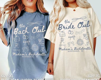 May include: Two oversized t-shirts with a bachelorette party theme. The blue jean shirt has white text that says "the Bach Club" and "Madison's Bachelorette Scottsdale, AZ". The ivory shirt has blue text that says "the Bride Club" and "Madison's Bachelorette Scottsdale, AZ".