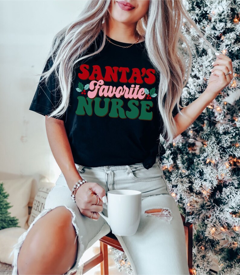 Puede incluir: Una mujer con una camiseta negra con el texto "Santa's Favorite Nurse" en letras rojas, verdes y rosas. Lleva una taza blanca y unos vaqueros azules claros.