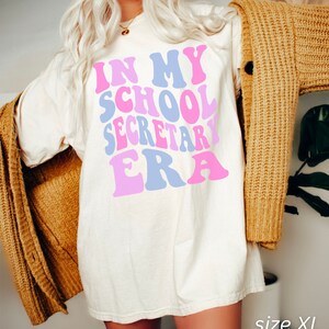 Peut inclure: Un t-shirt blanc surdimensionn&eacute; avec le texte "In My School Secretary Era" en rose, bleu et rose clair en police r&eacute;tro. Le t-shirt est pr&eacute;sent&eacute; sur un mannequin portant un cardigan marron. Le texte "size XL IVORY" est affich&eacute; en bas de l'image.