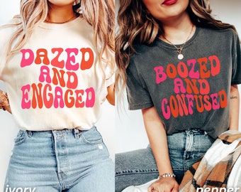 May include: Two women wearing t-shirts with retro-style text. The shirt on the left is ivory with the text "Dazed and Engaged" in pink and orange. The shirt on the right is grey with the text "Boozed and Confused" in pink and orange.