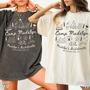 May include: Two short-sleeved t-shirts, one dark gray and one ivory, feature a camping-themed graphic. The design includes a tent, campfire, and text that reads "Camp Madelyn" and "Madelyn's Bachelorette, Breckenridge, CO."