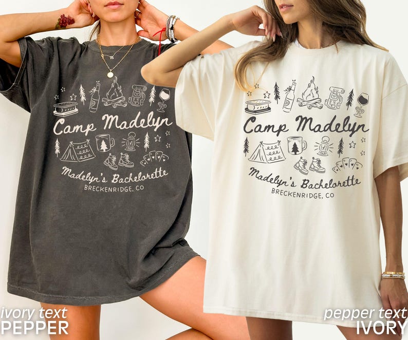 May include: Two short-sleeved t-shirts, one dark gray and one ivory, feature a camping-themed graphic. The design includes a tent, campfire, and text that reads "Camp Madelyn" and "Madelyn's Bachelorette, Breckenridge, CO."