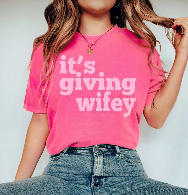 May include: A pink t-shirt with the text "it's giving wifey" in a bold, pink font.