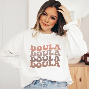 Op de afbeelding: Wit sweatshirt met het woord "Doula" vier keer herhaald in een gestapelde, retro-stijl lettertype. De tekst is in tinten roze en bruin.