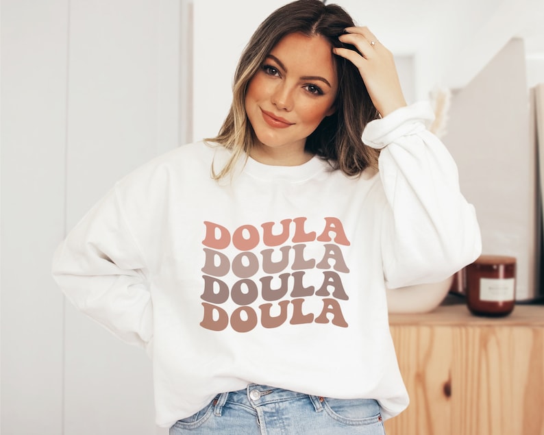 Op de afbeelding: Wit sweatshirt met het woord "Doula" vier keer herhaald in een gestapelde, retro-stijl lettertype. De tekst is in tinten roze en bruin.