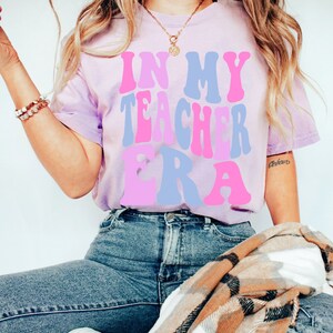 May include: A light purple t-shirt with the text "In My Teacher Era" in pink and blue letters. The shirt is being worn by a person with long blonde hair and is sitting down.