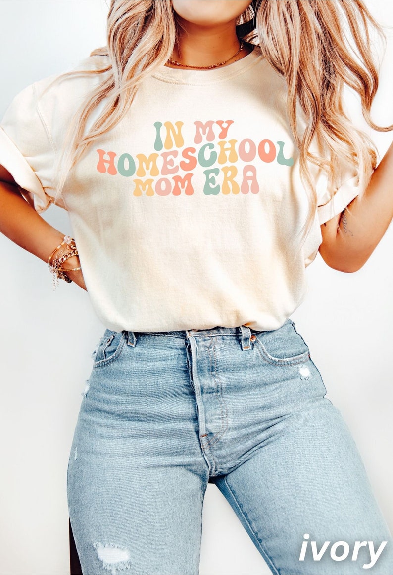 May include: Cream-coloured t-shirt featuring the text "IN MY HOMESCHOOL MOM ERA" in a retro font. The shirt is worn with light blue denim jeans. The style is casual and comfortable, suitable for everyday wear.