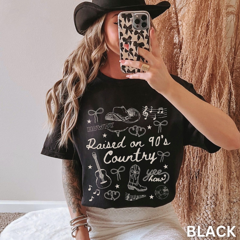 May include: Black t-shirt with white graphic text and illustrations. The text reads "Raised on 90's Country" with cowboy-themed drawings. The shirt also features the word "Howdy" and the phrase "yee haw".