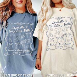 May include: Two XL t-shirts, one blue and one ivory, with a decorative frame design. The text reads "Seashells & Wedding Bells" and "Emma's Bachelorette Siesta Key, FL". The design includes seashells, palm trees, and cocktail illustrations.