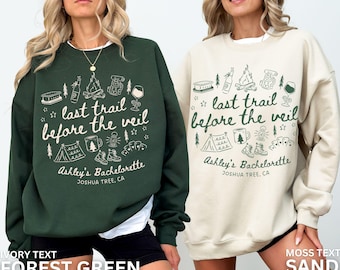 May include: Two sweatshirts, one forest green and one sand-coloured, display the text "last trail before the veil" with camping-themed illustrations. The text "Ashley's Bachelorette, Joshua Tree, CA" is also included.