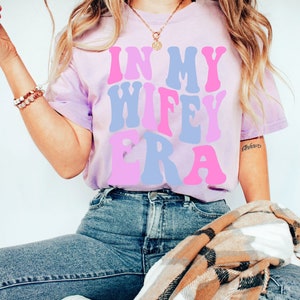 May include: A light purple t-shirt with the text "In My Wife Era" in pink and blue letters. The shirt is being worn by a person and is available in size S.