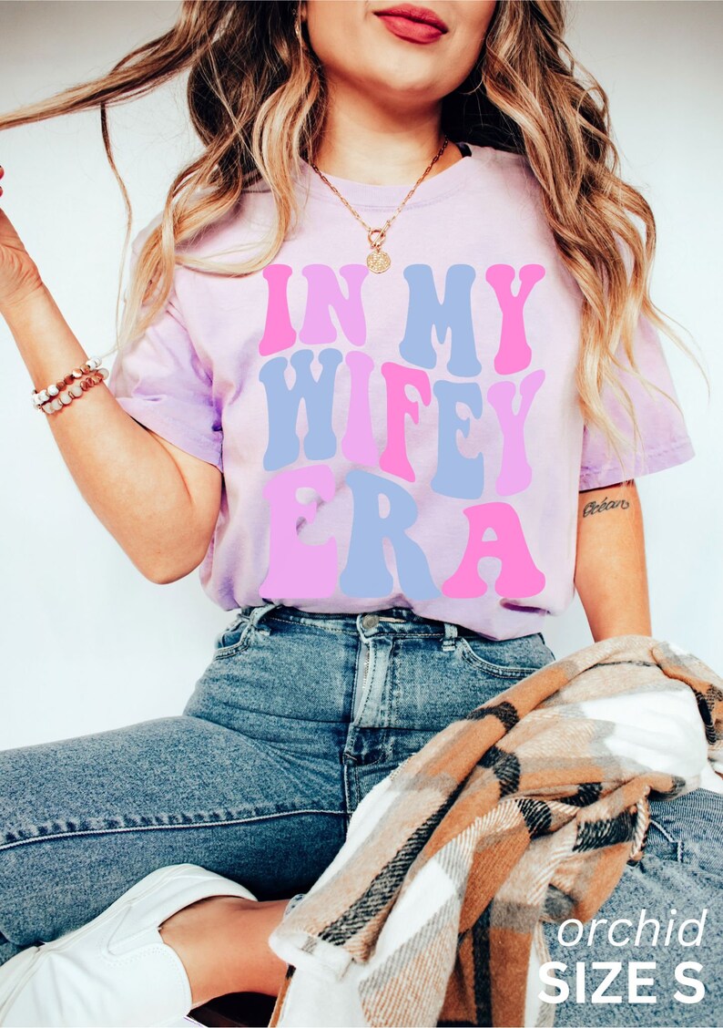 May include: A light purple t-shirt with the text "In My Wife Era" in pink and blue letters. The shirt is being worn by a person and is available in size S.