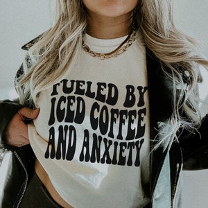 Peut inclure: T-shirt ivoire avec l'inscription noire "Fueled by Iced Coffee and Anxiety" dans une typographie r&eacute;tro. Le t-shirt de taille S est port&eacute; avec une veste en cuir noire et un collier cha&icirc;ne dor&eacute;.