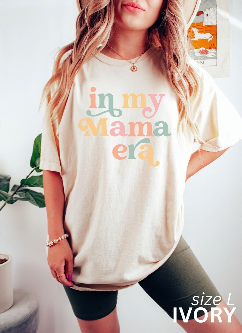 Op de afbeelding: Een wit T-shirt met de tekst "in my mama era" in kleurrijke retro letters. Het shirt wordt gedragen door een persoon en is maat L. Het shirt is ivoor.