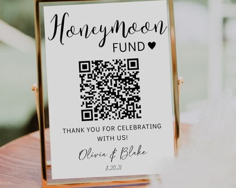 May include: White sign with black text that reads "Honeymoon Fund" with a black heart symbol. A black and white QR code is below the text. The text "Thank you for celebrating with us!" is below the QR code. The names "Olivia & Blake" and the date "8.20.21" are below the text. The sign is in a gold frame.